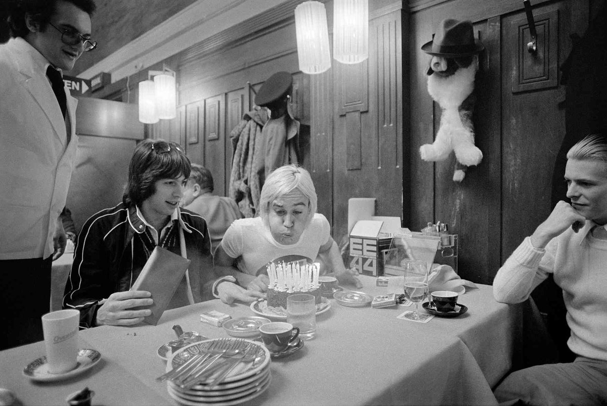 Iggy Pop blows out the candles on his birthday cake as David Bowie and tour manager Pat Gibbons look on at a restaurant in Basel Switzerland 1976